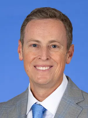 Smiling man in gray plaid suit jacket and blue tie against a solid blue background, professional portrait.