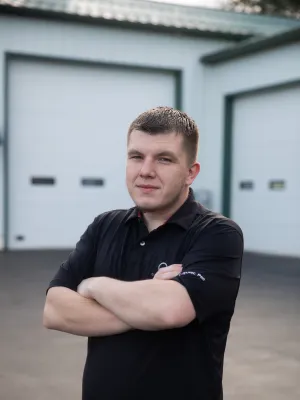 A confident young man in a black polo shirt, standing with arms crossed in front of a garage.
