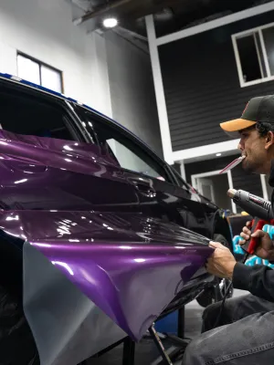 A professional applying purple vinyl wrap to a blue car in an auto shop.