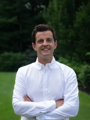 Smiling man in a white shirt stands confidently outdoors with arms crossed in a lush green setting