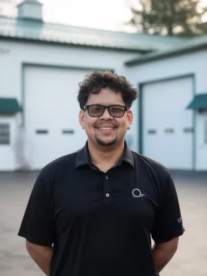 Smiling man in a black polo shirt stands outside a garage, showcasing friendly demeanor and professional attire.