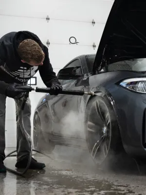 Worker detailing a luxury car with a pressure washer in a professional garage setting.