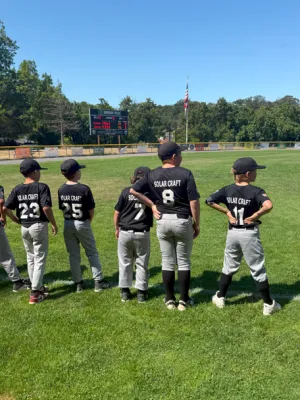 Youth baseball players in Solar Craft uniforms stand in a row on a grassy field with scoreboard and trees behind.
