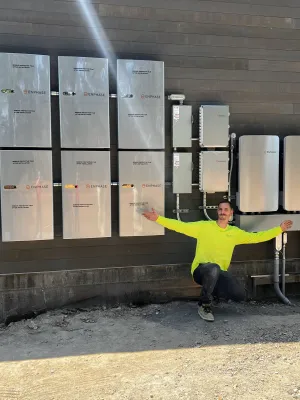 Man in yellow shirt kneeling next to multiple Enphase solar energy system components mounted on a brown exterior wall.