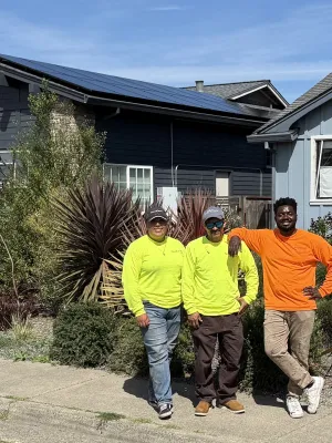 Three workers in bright shirts standing on a sidewalk in front of houses with solar panels on the roof under a clear blue sky