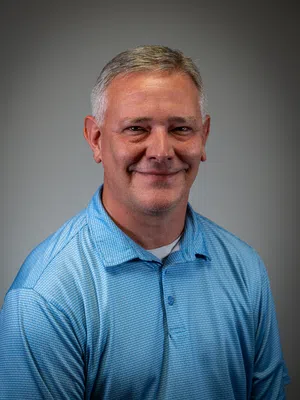 Portrait of a middle-aged man with short gray hair wearing a blue polo shirt smiling against a gray background