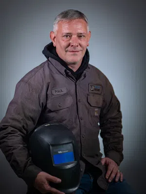 Mid-aged man in workwear holding a welding helmet, posing confidently against a neutral background.