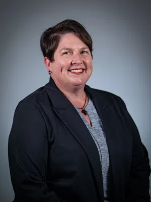 Smiling woman with short brown hair wearing a floral patterned dark blouse on a gray background