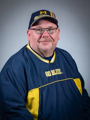 Middle-aged man wearing glasses, a Michigan Big House cap, and a Go Blue jacket with navy and yellow colors.
