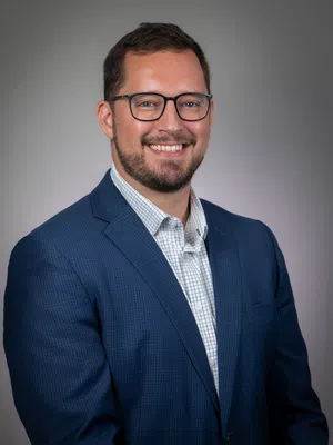 Smiling man with beard and glasses wearing blue blazer and checkered shirt on gray background