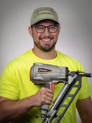Smiling man in glasses and cap holds Paslode nail gun wearing bright yellow shirt against gray background