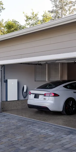 White Tesla Model X parked in a suburban garage with a Tesla Powerwall on the wall and stone pillars.