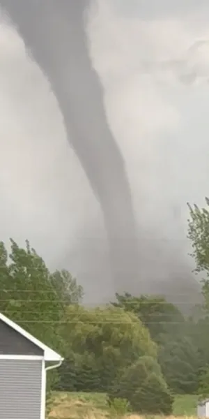 Tornado funnel touching down near houses and trees under cloudy sky during a storm.