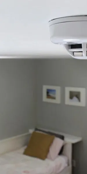 White smoke detector mounted on ceiling in a modern bedroom with gray walls and a bed.