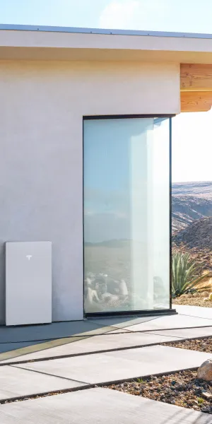 Modern house exterior with solar battery unit, wooden bench, desert plants, and mountainous background under clear sky.