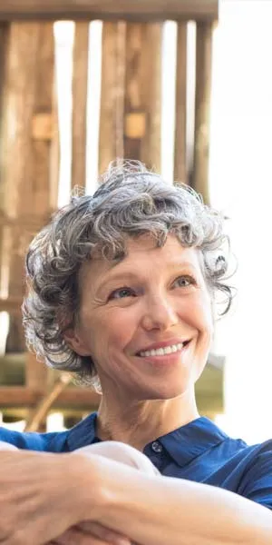 Smiling mature woman with curly gray hair sitting with arms crossed, looking away thoughtfully indoors.