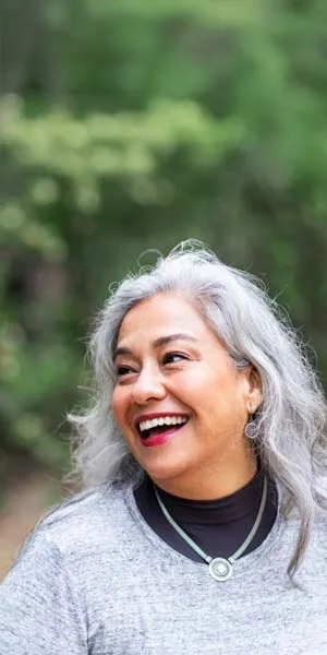 Smiling older woman with gray hair outdoors wearing a gray sweater and necklace against blurred green background