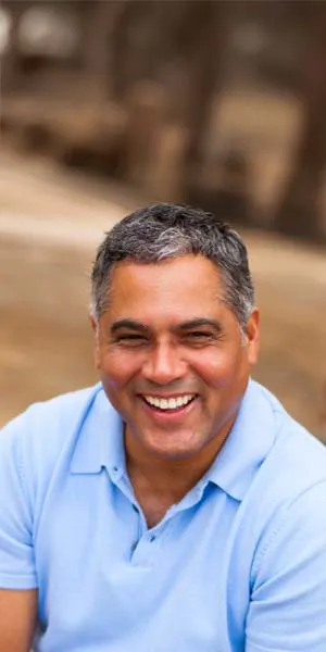 Middle-aged man with salt-and-pepper hair smiling warmly, wearing a light blue polo shirt outdoors.