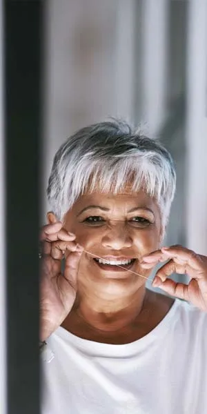 Smiling mature woman with short gray hair flossing her teeth indoors wearing a white top.