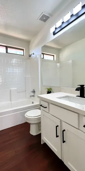 Modern white bathroom with bathtub, toilet, vanity with black faucet, and large mirror, featuring wood flooring.