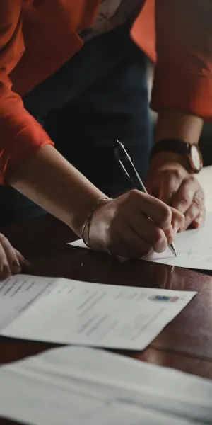 Person in red jacket signing documents on a wooden table with another person observing.