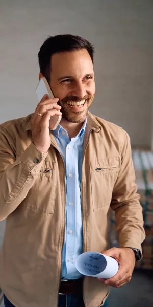 Smiling man in beige jacket talking on phone holding blueprint with worker and construction plans in background
