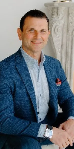 Man in blue suit sitting on beige chair smiling in a stylish room with books and ornate fireplace.