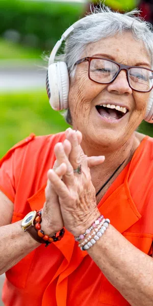 Happy elderly woman in orange shirt enjoying music with headphones outdoors in a green park setting.