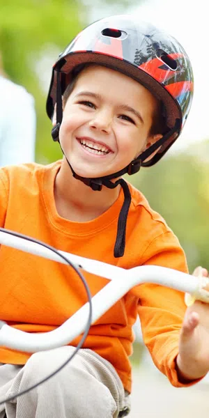 Smiling boy in orange shirt wearing helmet rides bike outdoors with woman standing behind in green park.