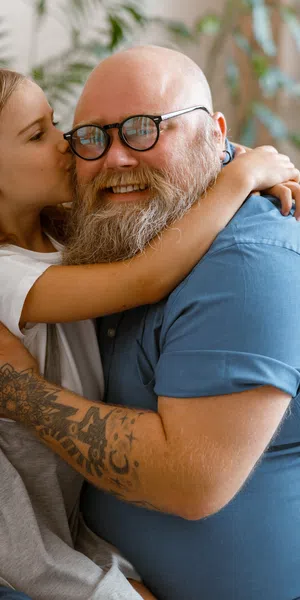 Smiling bearded man with tattoos hugging and being kissed by a young girl in a cozy home setting.