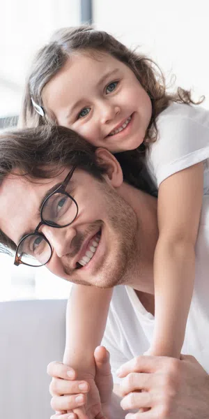 Smiling father wearing glasses carries happy daughter on his back in cozy living room with large window.