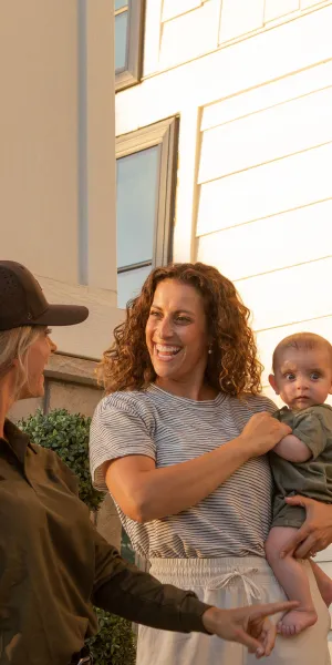 Woman holding baby greets cleaner with duster outside modern home during daytime