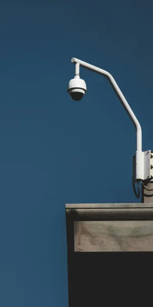White surveillance camera mounted on a beige building corner against a clear blue sky.