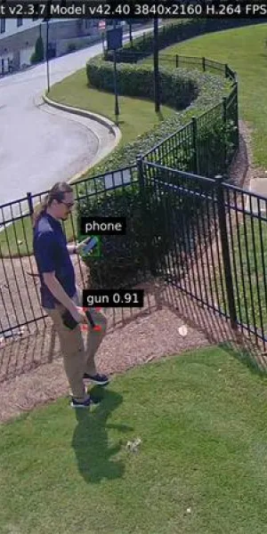 Person standing near a fence holding an object detected as a gun by AI with phone also detected in hand.