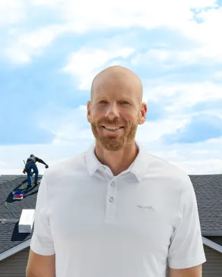 Smiling man in white polo stands in front of roofers installing shingles on a house under a partly cloudy sky