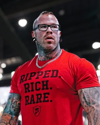 Muscular tattooed man wearing glasses and red shirt with RIPPED RICH RARE text in a bright indoor setting.