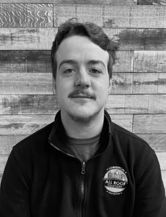 Black and white portrait of a young man with mustache wearing All Roof Solutions jacket against wooden wall.