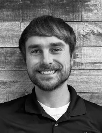 Smiling man with beard and short hair wearing a black polo shirt standing in front of wooden wall