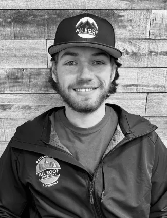 Smiling man wearing All Roof Solutions cap and jacket against wooden plank background in black and white photo