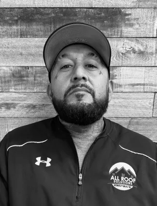 Bearded man wearing a baseball cap and All Roof Solutions jacket in front of a wooden wall.