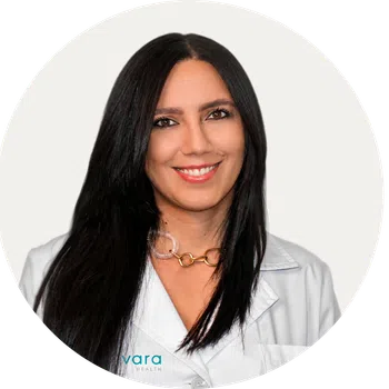 Portrait of a smiling female healthcare professional with long black hair wearing a white coat and gold necklace.