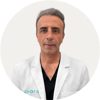 Middle-aged male doctor wearing a white coat and black scrubs with gray hair against a plain background.