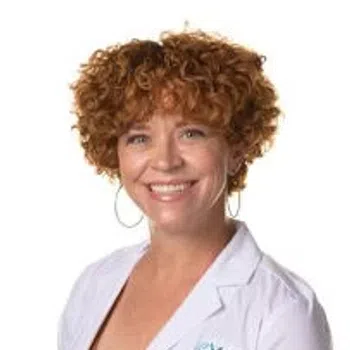 Smiling woman with curly red hair wearing a white Evora medical coat and hoop earrings against white background