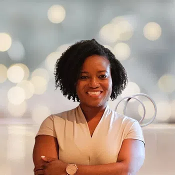 Smiling woman with curly hair and crossed arms wearing a beige top and watch against a blurred light background.