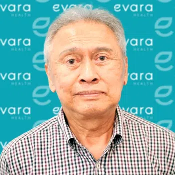 Older man in a checked shirt stands in front of a blue backdrop with Evara Health logos.