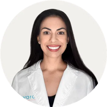 Smiling woman with long dark hair wearing a white lab coat against a plain light background