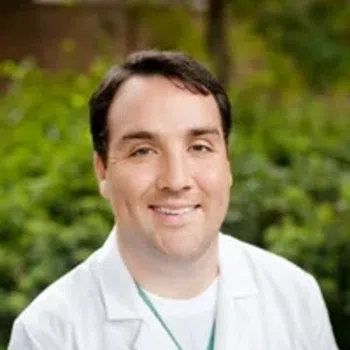 Smiling male doctor wearing a white lab coat with a blurred green outdoor background.