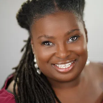 Close-up portrait of a smiling Black woman with long dreadlocks wearing pearl earrings and a burgundy top.