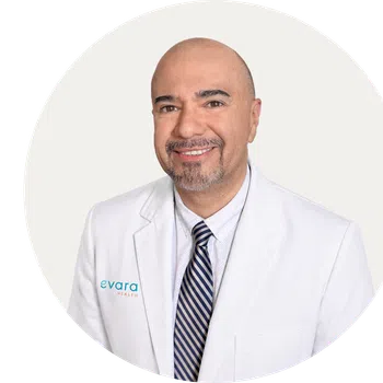 Smiling bald man with goatee wearing a navy blazer, blue shirt, and red-striped tie against a beige background.