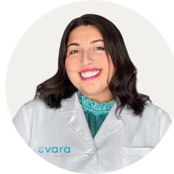 Smiling woman in white medical coat with teal blouse, Evara Health logo visible on coat pocket.